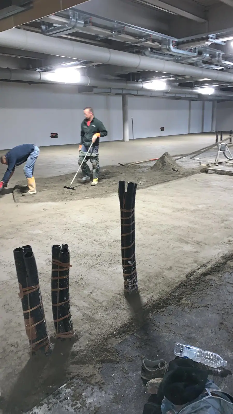 Due operatori lavorano alla posa del massetto in un ampio locale, attorno alle tubazioni verticali.