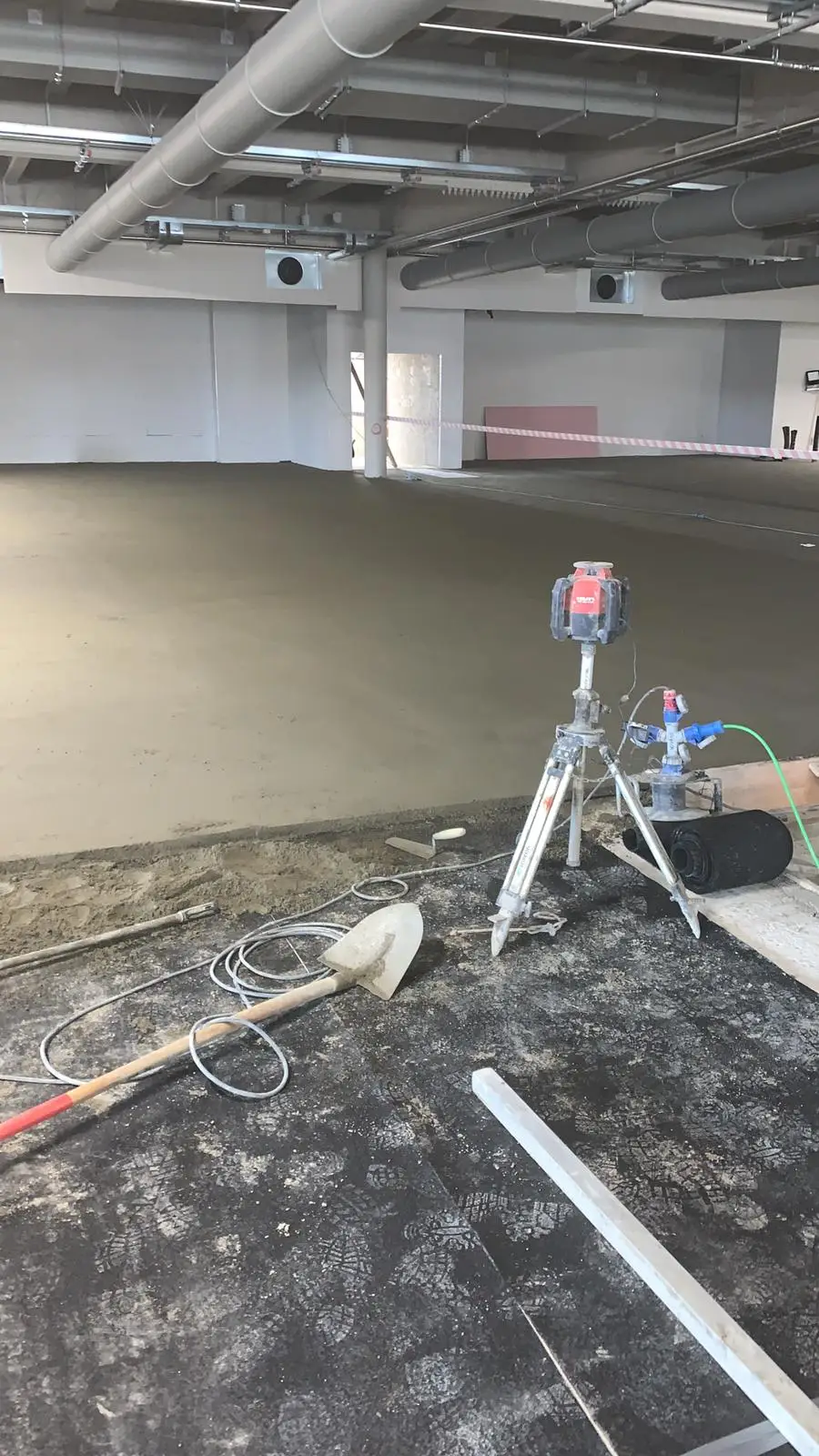 Strumento su treppiede e tubazioni davanti al massetto di un ampio locale con impianti a soffitto.